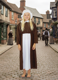 A young girl in a brown dress and white bonnet stands on a cobblestone street, surrounded by traditional English buildings and other people.